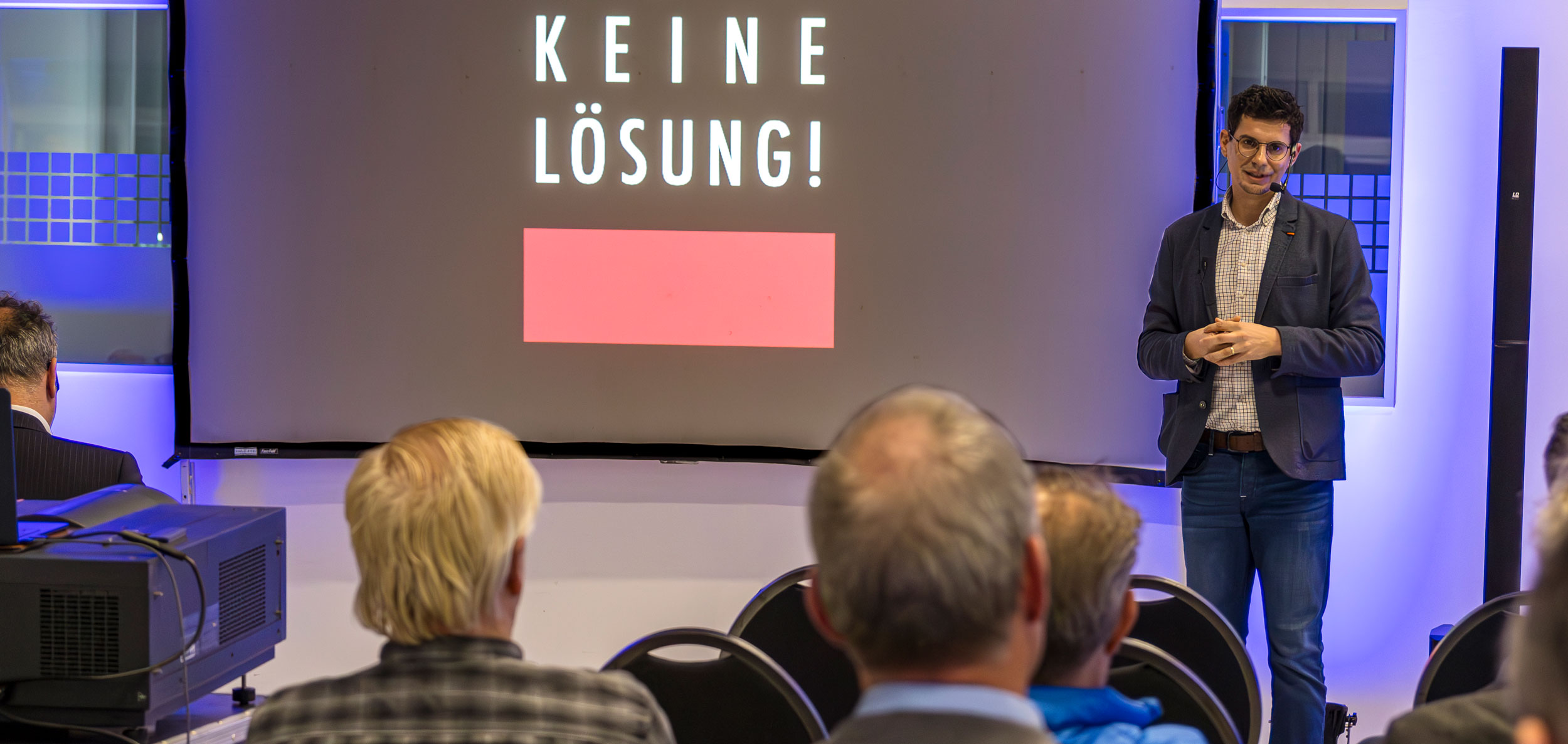 Sebastian Kupfer steht vor einigen Zuhörern. Im Hintergrund ist eine Präsentation zu sehen mit dem Titel "Dagegen sein ist keine Lösung!"